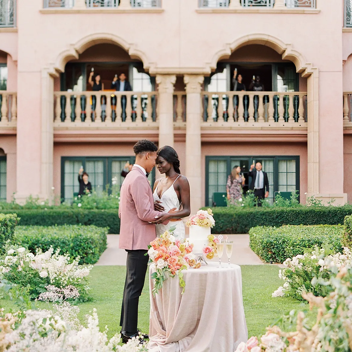 2022 Weddings Survey: A couple embracing with their wedding guests cheering from the building behind them.