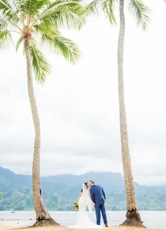 A wedding couple kissing in between two palm trees overlooking the Pacific Ocean in Princeville, Hawaii, photographed by Photography By Hanakapi’ai. 