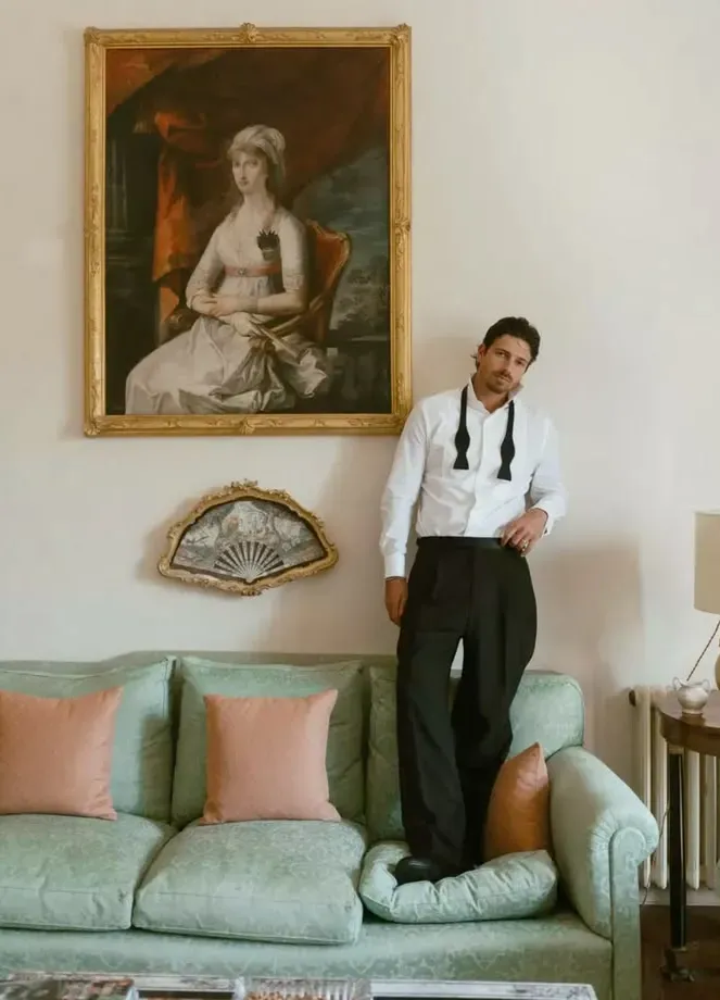 Groom in a white shirt and black trousers with a long black ribbon tie, standing in a green-and-gold sitting room. Ribbon tie without a jacket.