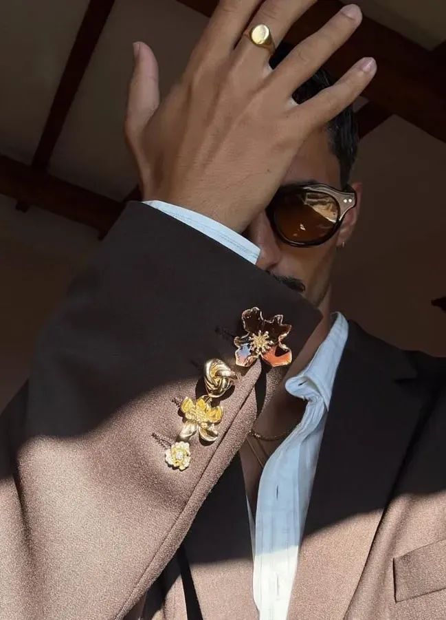 Tight crop of a tan lapel carrying a cluster of enamel and gold floral brooches, with an open pale blue shirt and a gold signet ring visible. 