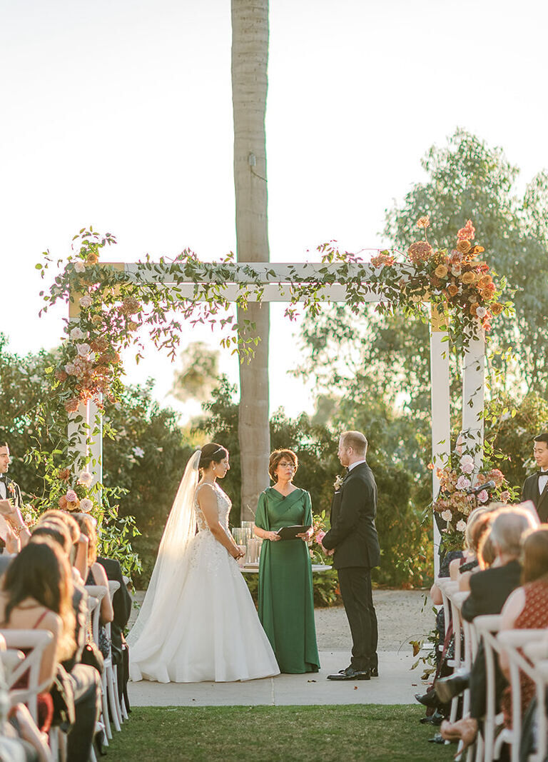 A couple stands under a white arch decorated with earth tone flowers at their outdoor wedding.