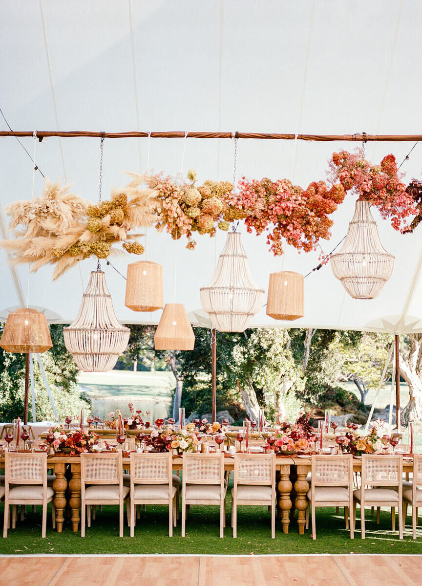 An earth tone wedding reception in a tent boasts an ombre floral installation, hanging light fixtures, and colorful floral arrangements atop wood tables set with wood chairs.