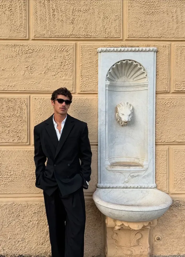 Groom in a relaxed black suit with an open collar, no tie, against an ochre wall with a stone fountain. 