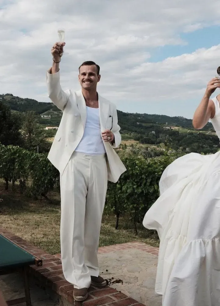 Groom in a relaxed ivory suit over a plain white tank, raising a glass at a vineyard, bride beside him in white.