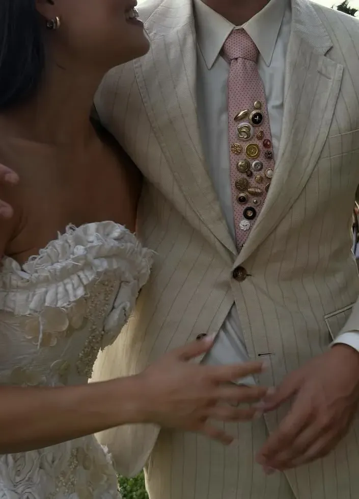 Cream pinstripe suit with a dense run of mismatched vintage buttons and pins down the placket, bride's hand at the waist. 