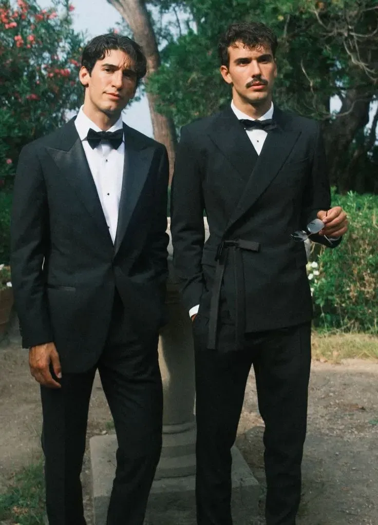 Two grooms outdoors in black tuxedos, one in a bow tie and one in a belted wrap jacket. 