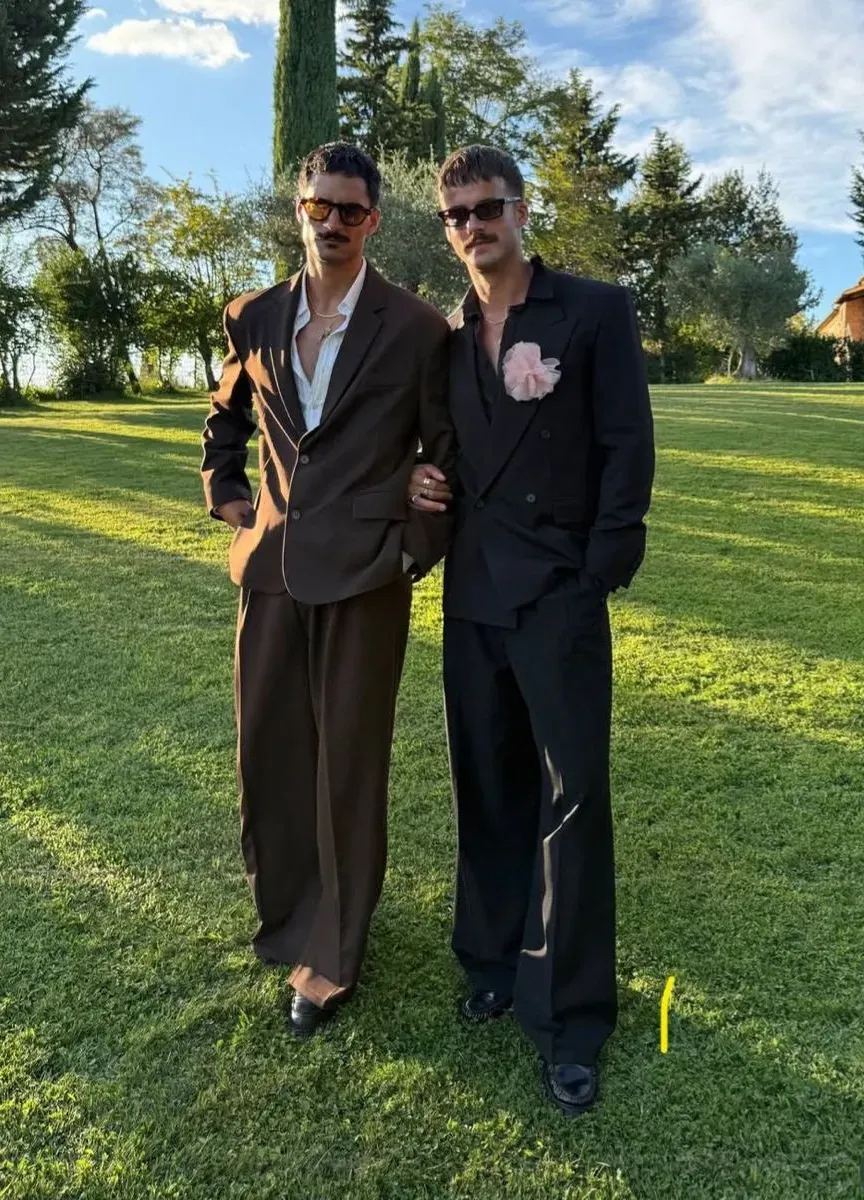 Two grooms on a lawn in relaxed brown and navy suits with wide trousers and contrast piping, one wearing a pale pink boutonnière. 