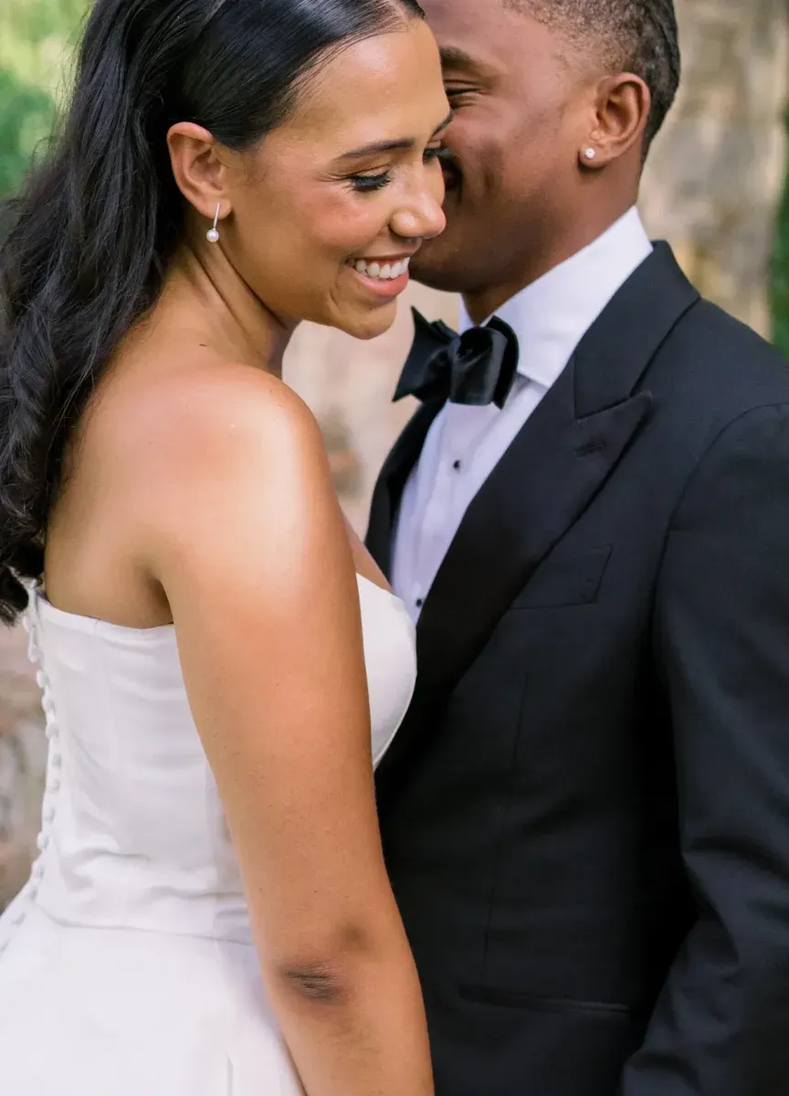 A bride in a strapless wedding gown smiling with a groom in a black tuxedo with a black bow tie smiling whispering in her ear. 