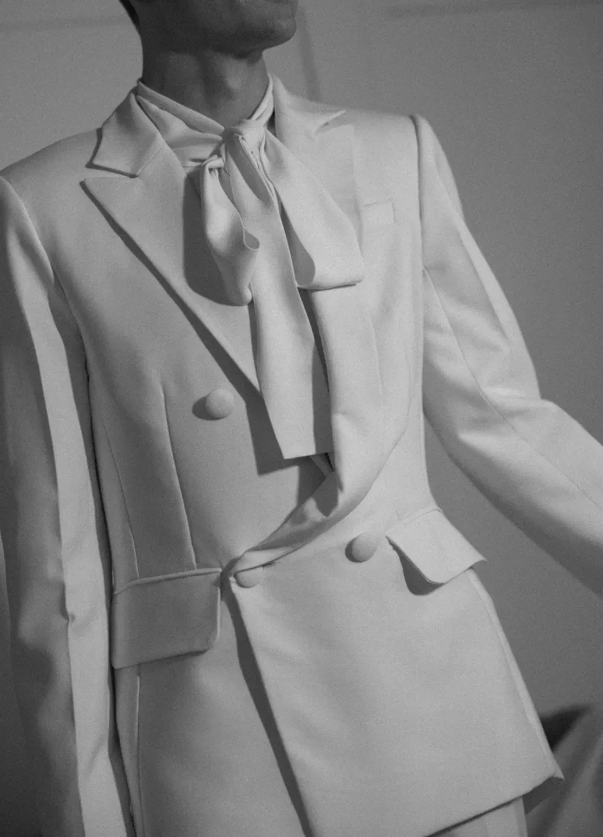 Studio-lit pale suit with a tonal scarf tie and covered buttons, tightly cropped at the neck and shoulder. 