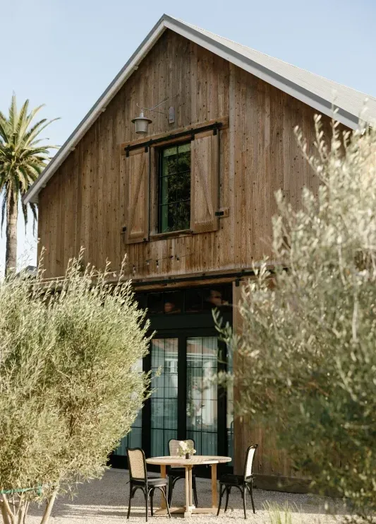 A modern-minimalist barn at a luxury resort in Los Olivos, California, with lush greenery and a small outdoor table in the foreground.