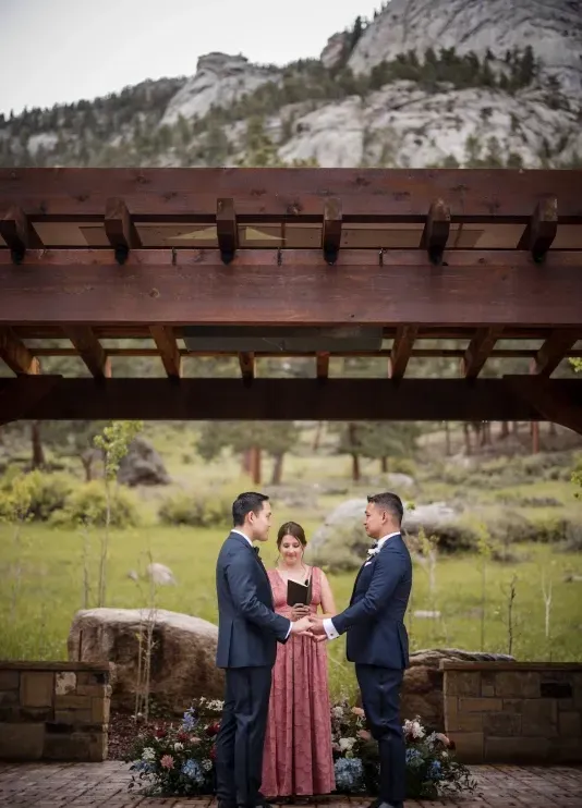Two grooms holding hands and their officiant reading from a booklet during their outdoor wedding ceremony at a mountainside chateau in Colorado, overlooking greenery and mountains in the distance.