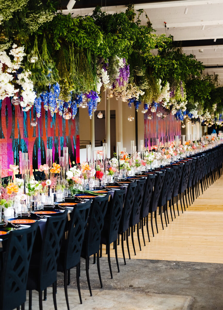 One long reception table was set in an industrial space with colorful art at a Boston couple's art museum wedding. Greenery and floral hang overhead, with modern black chairs balancing the design below.