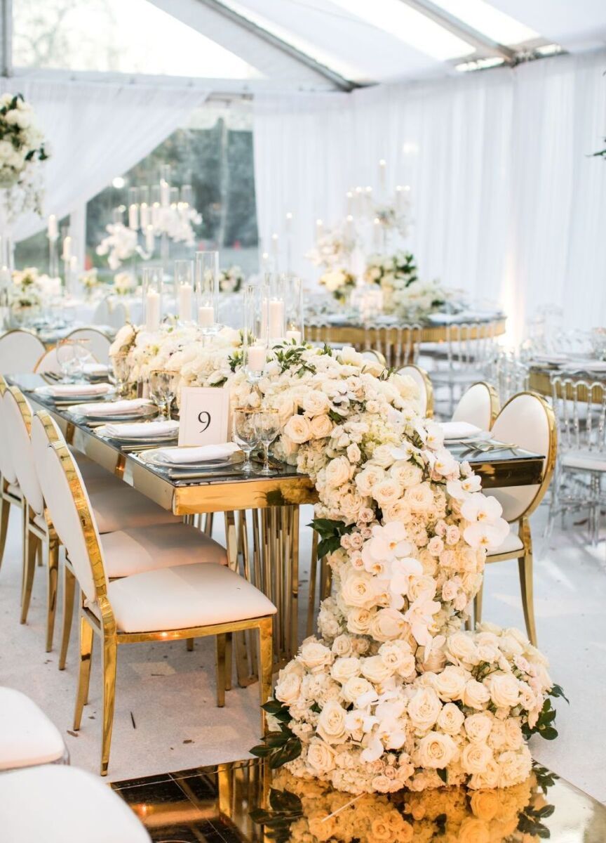 White and gold tented wedding reception featuring a long table with white floral runner cascading down on to the floor