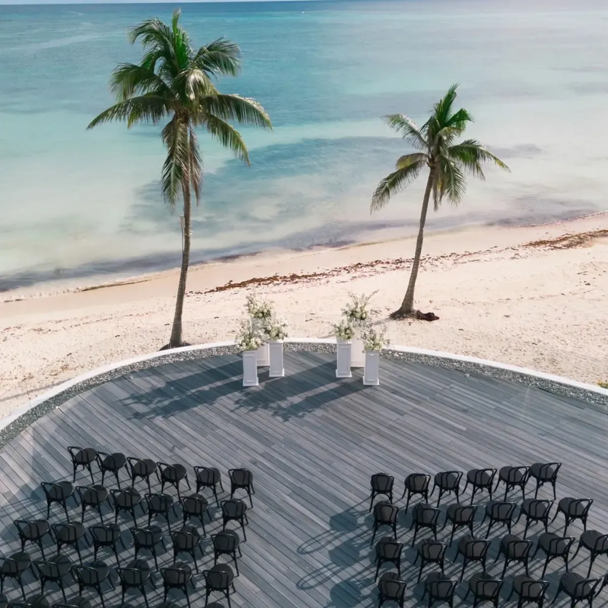 outdoor beachfront wedding ceremony setup with white floral arrangements and black chairs overlooking turquoise water