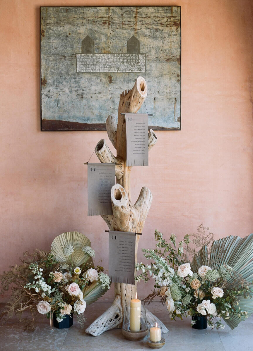 An escort card alternative, with fringed leather seating charts hanging on driftwood, surrounded by floral arrangements and candles.