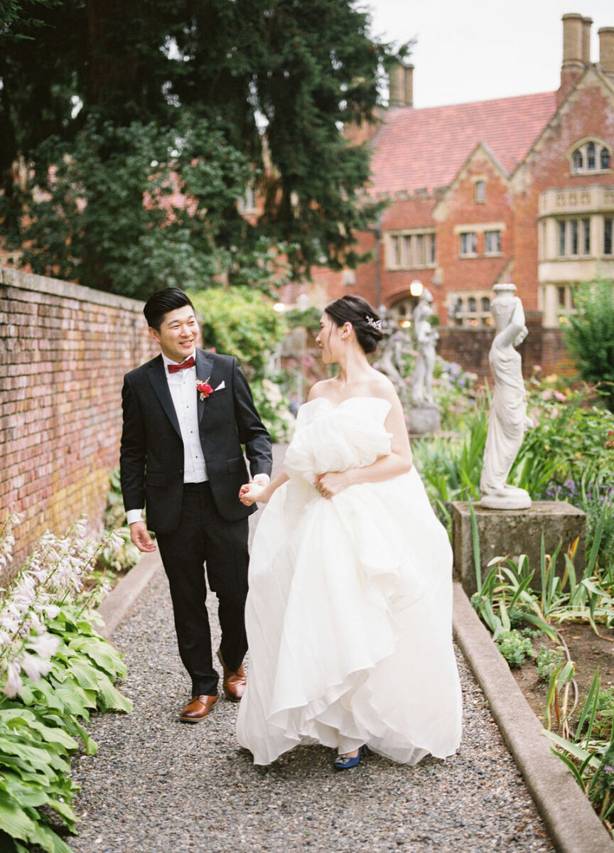 Castle Wedding Venues: A wedding couple holding hands and smiling while walking on a pathway lined with greenery, old statues, and a red brick castle behind them.