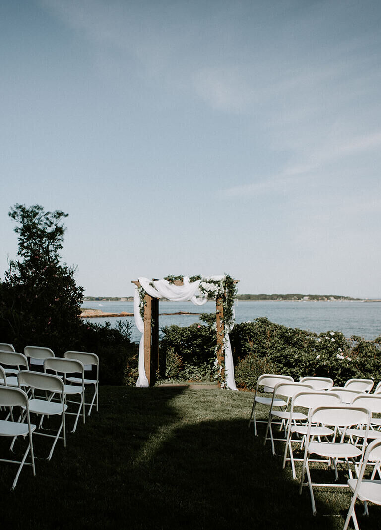 Castle Wedding Venues: A ceremony setup with a wooden arch overlooking the water at a castle wedding venue in Gloucester, Massachusetts.