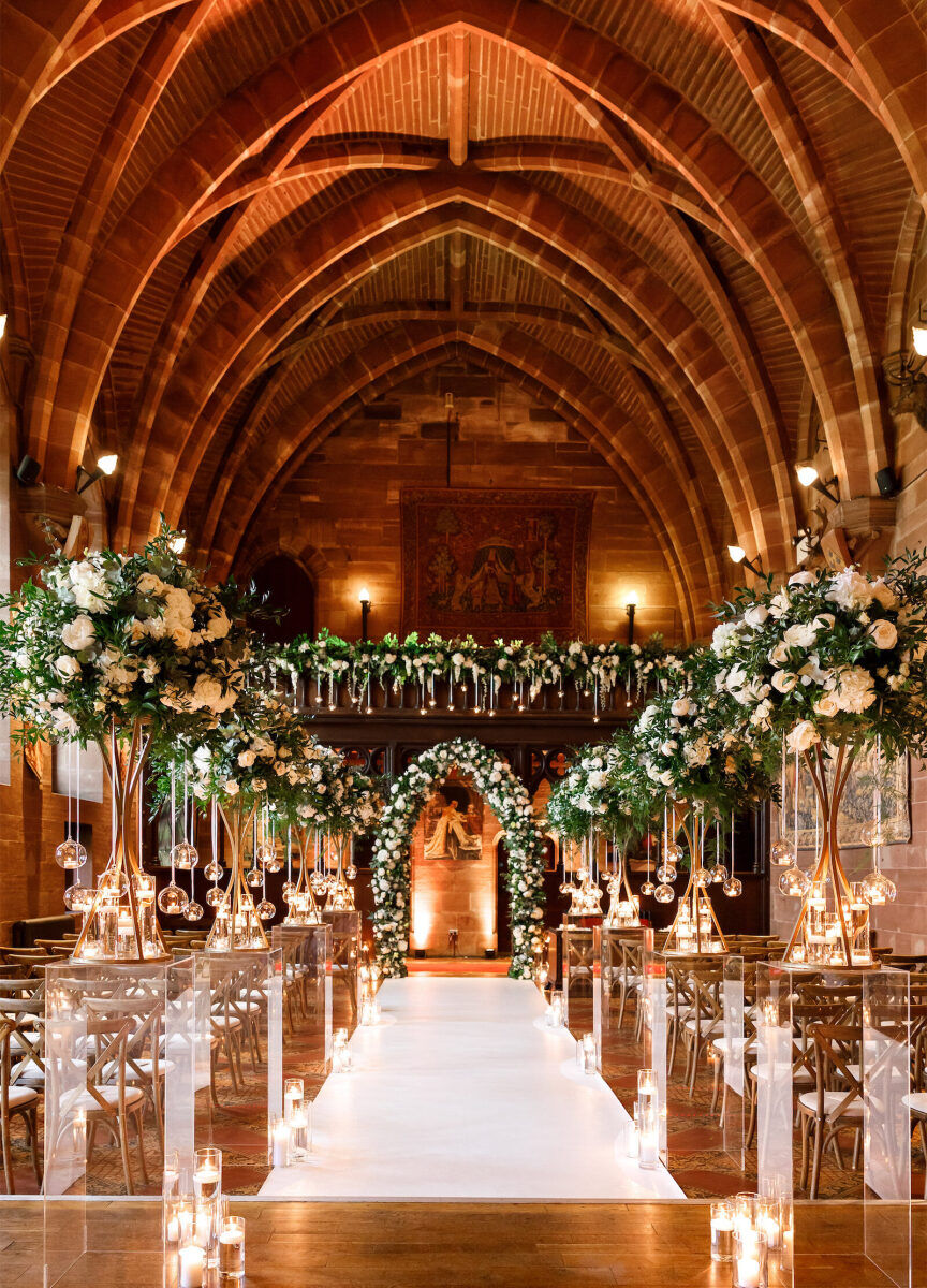 Castle Wedding Venues: An English castle's interior with an elegant ceremony setup, with wooden chairs and a candle-lit aisle, tall floral arrangements and a green and white arch at the end.