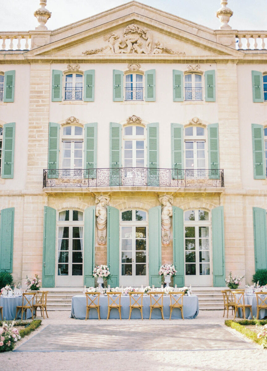 Castle Wedding Venues: An outdoor reception setup with long tables and white floral arrangements outside of a castle in France with green shutters.