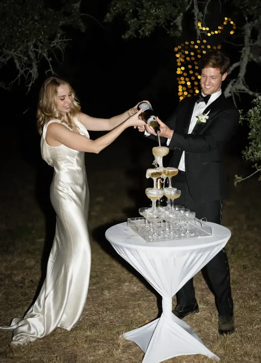 A couple pouring a bottle of champagne onto a tower of champagne flutes, with the bride in a silk floor-length gown and a groom in a black tuxedo.