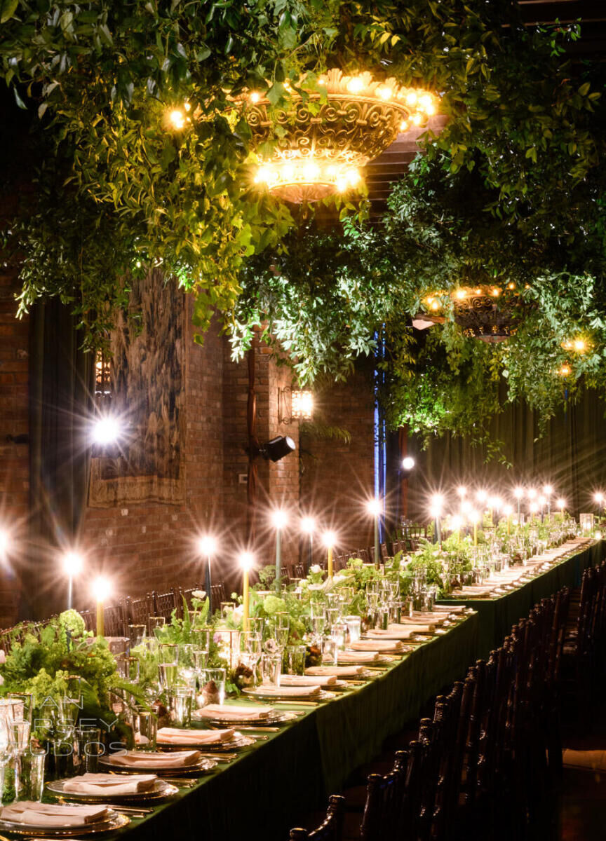 Ceiling Wedding Decor: A dark reception setting with a large light fixture covered in greenery and a long rectangular table with long-stemmed candles.