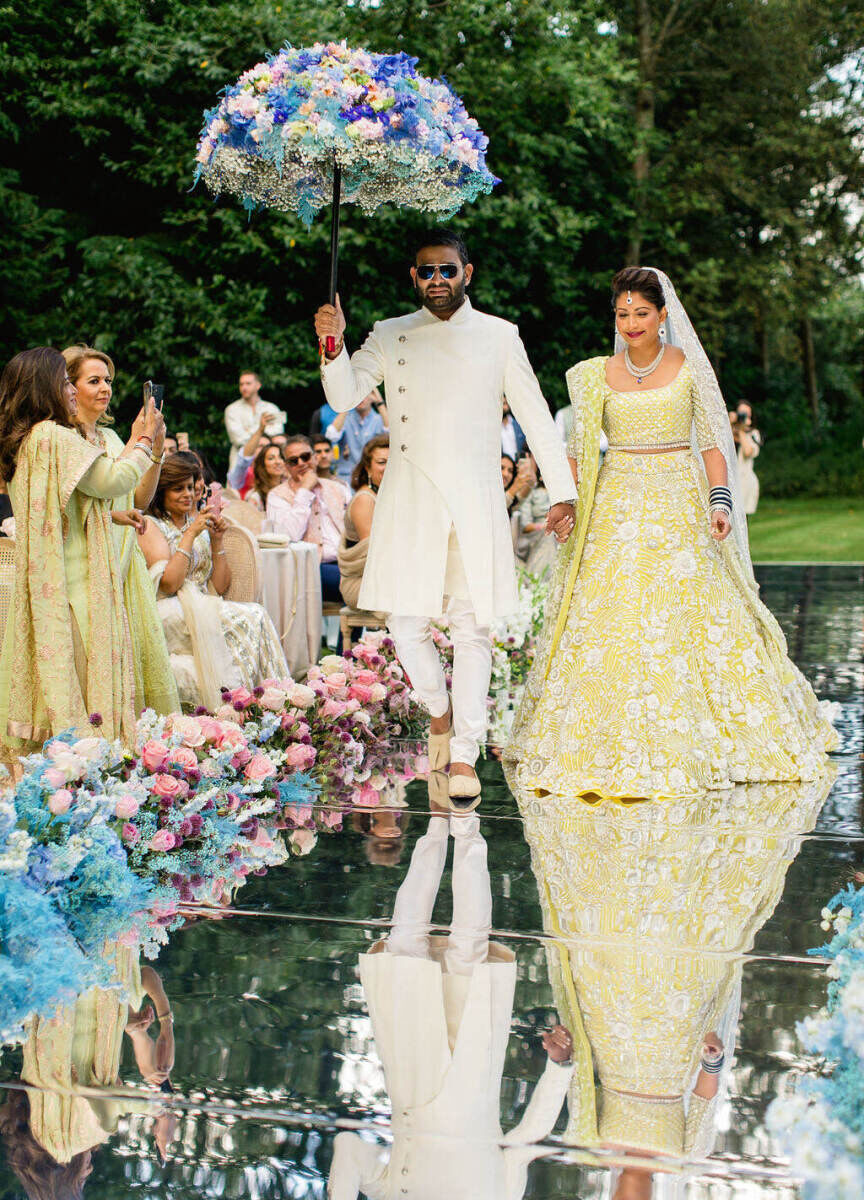 A bride is escorted down the aisle by her brother, who holds a flower-covered parasol to symbolize family and love, during her colorful countryside wedding ceremony.