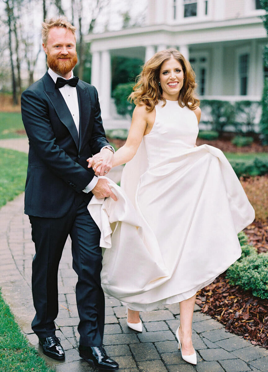 A groom and bride smile as they walk hand in hand, with him holding the bottom of her dress, outside of their North Carolina wedding venue.