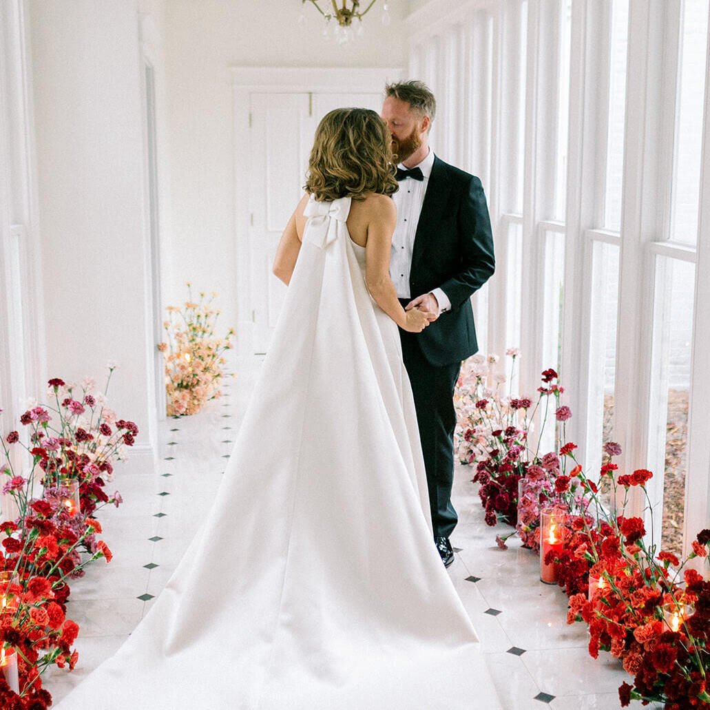 A bride and groom hold hands in a hallway decorated with a gradient of colorful flowers and glowing candles.