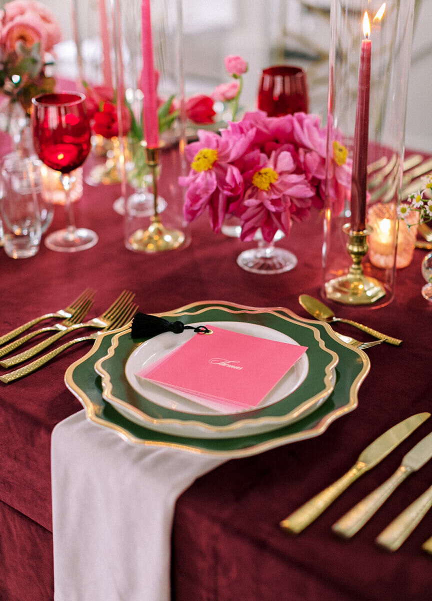 A placesetting, with multiple pieces of gold flatware for the multi-meal dinner reception, flank a stack of fluted green plates and a pink menu with a tassel on it.