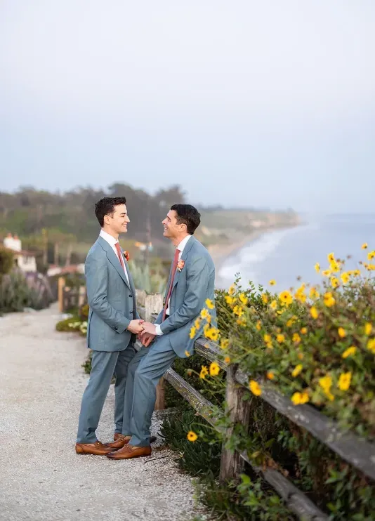 Two grooms smiling at each other on a yellow flower-lined path overlooking the ocean in Santa Barbara, California.