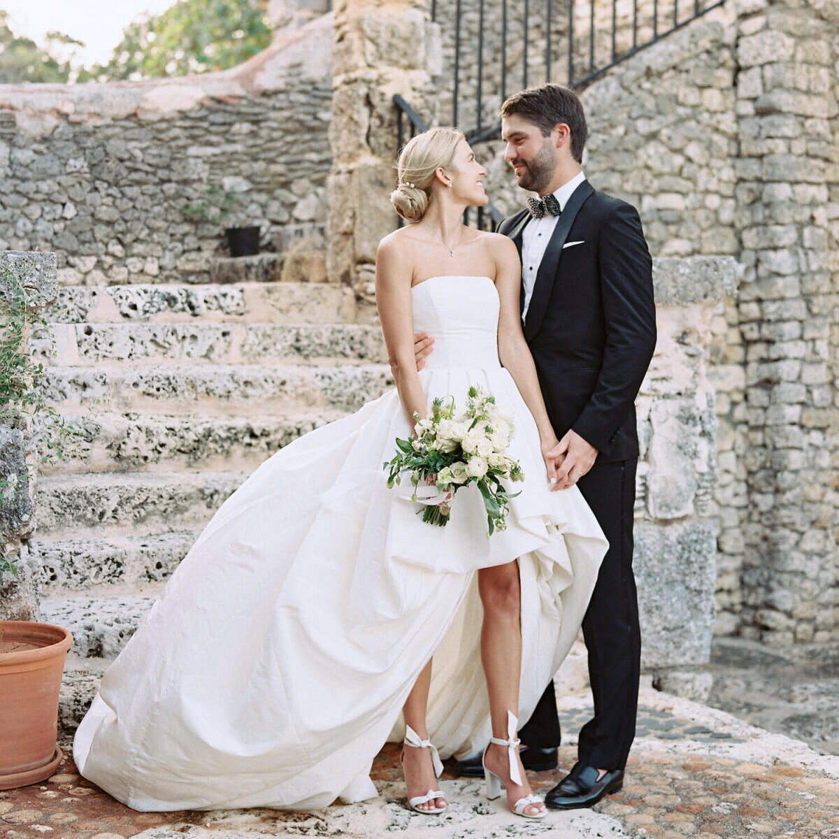 A bride and groom hold hands and lock eyes—she in a high-low dress and holding a bouquet of white flowers, and he in a classic tuxedo—during pre-ceremony portraits at their destination wedding in the Dominican Republic.