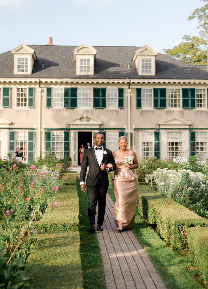 A groom and his mother walk down a manicured courtyard to his elevated garden wedding ceremony.