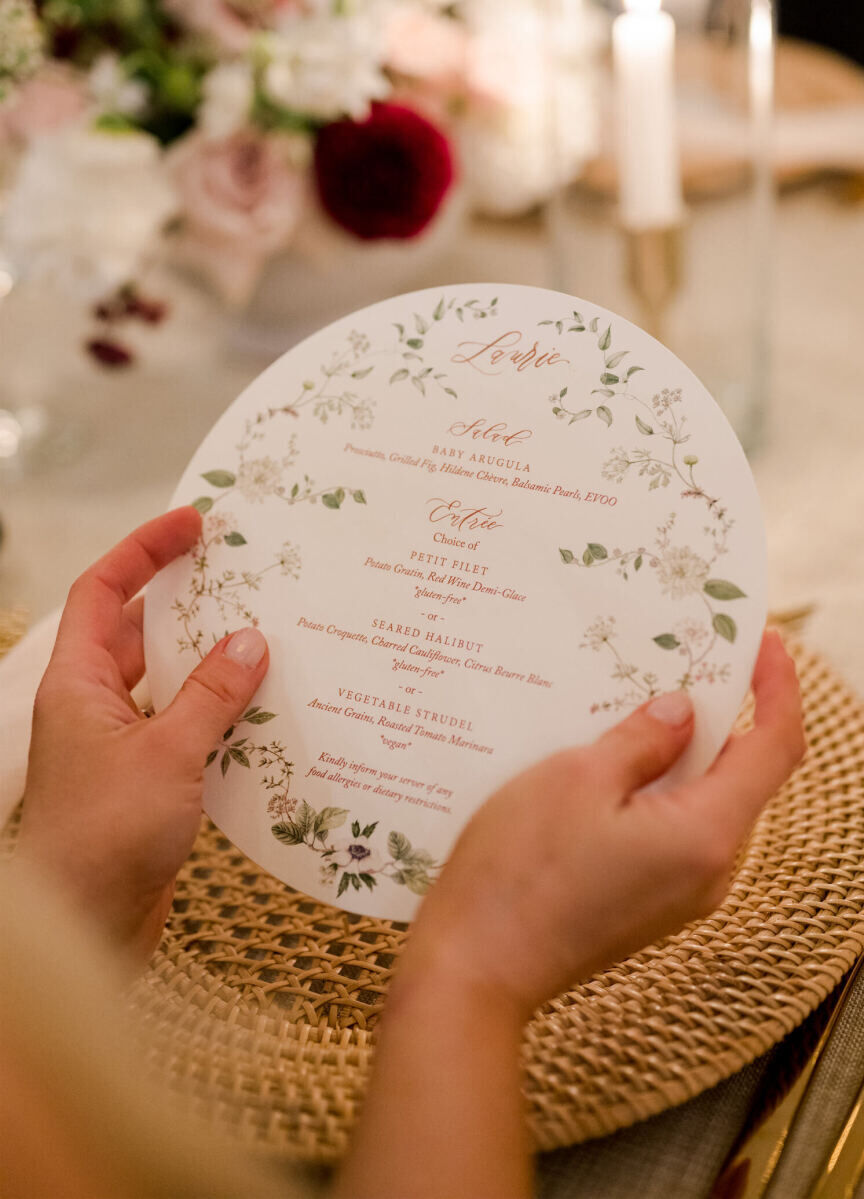 A guest holds a round menu decorated with floral illustrations and calligraphy at an elevated garden wedding.