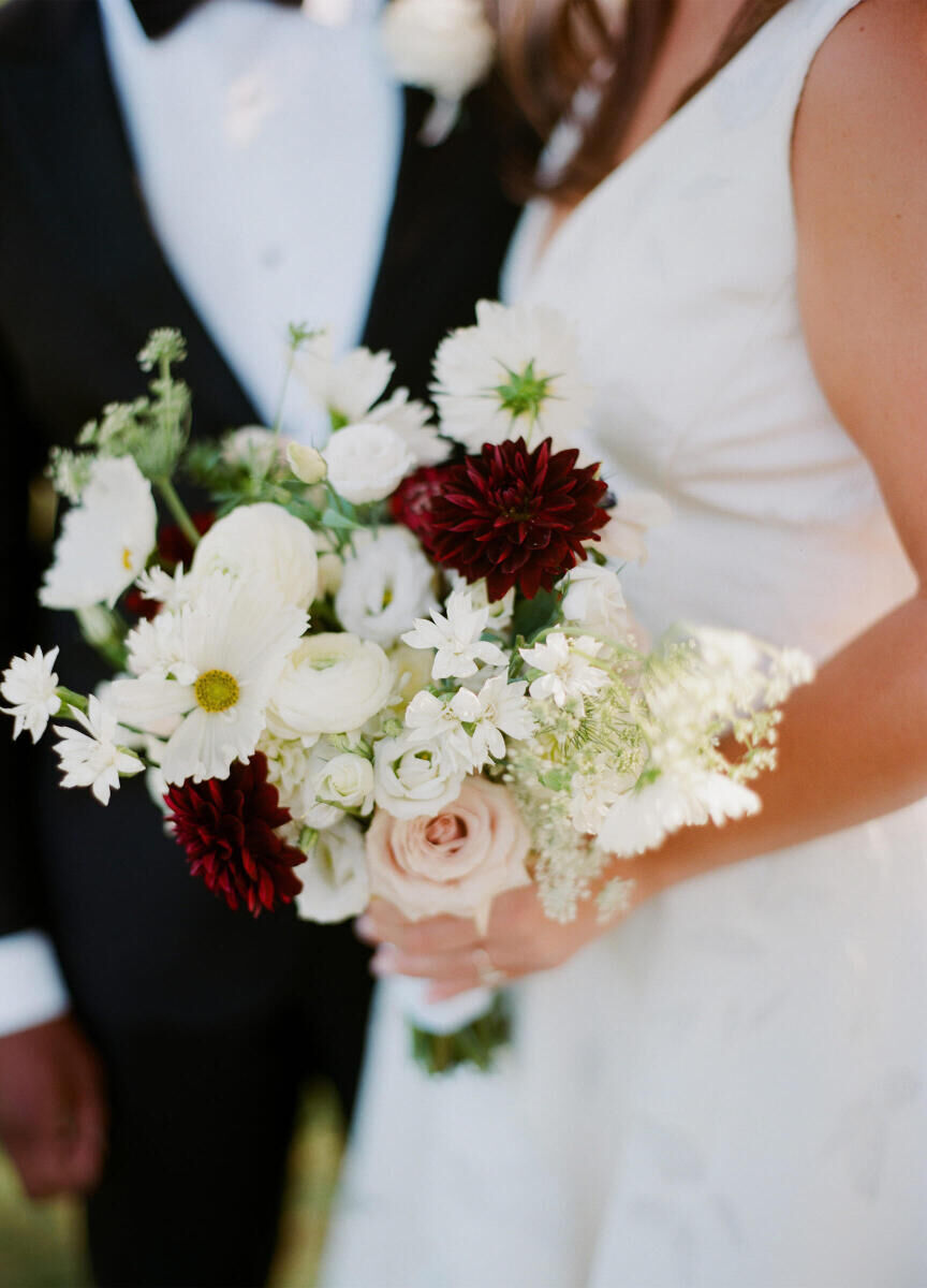 A bride holds a bouquet of mostly white flowers, with pops of soft pink and deep red, at her end-of-summer elevated garden wedding.