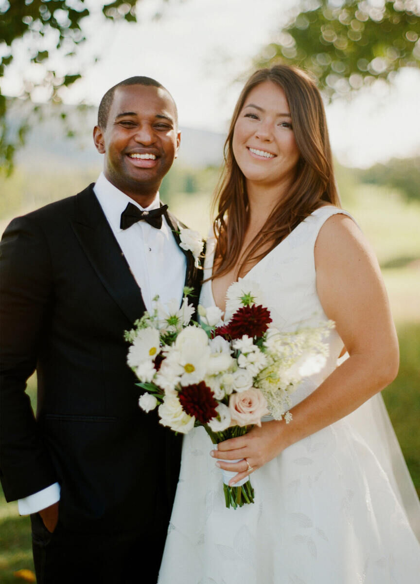 A groom and bride (holding a bouquet of white, pink, and burgundy blooms and sporting soft, loose waves in her hair) smile at their elevated garden wedding.