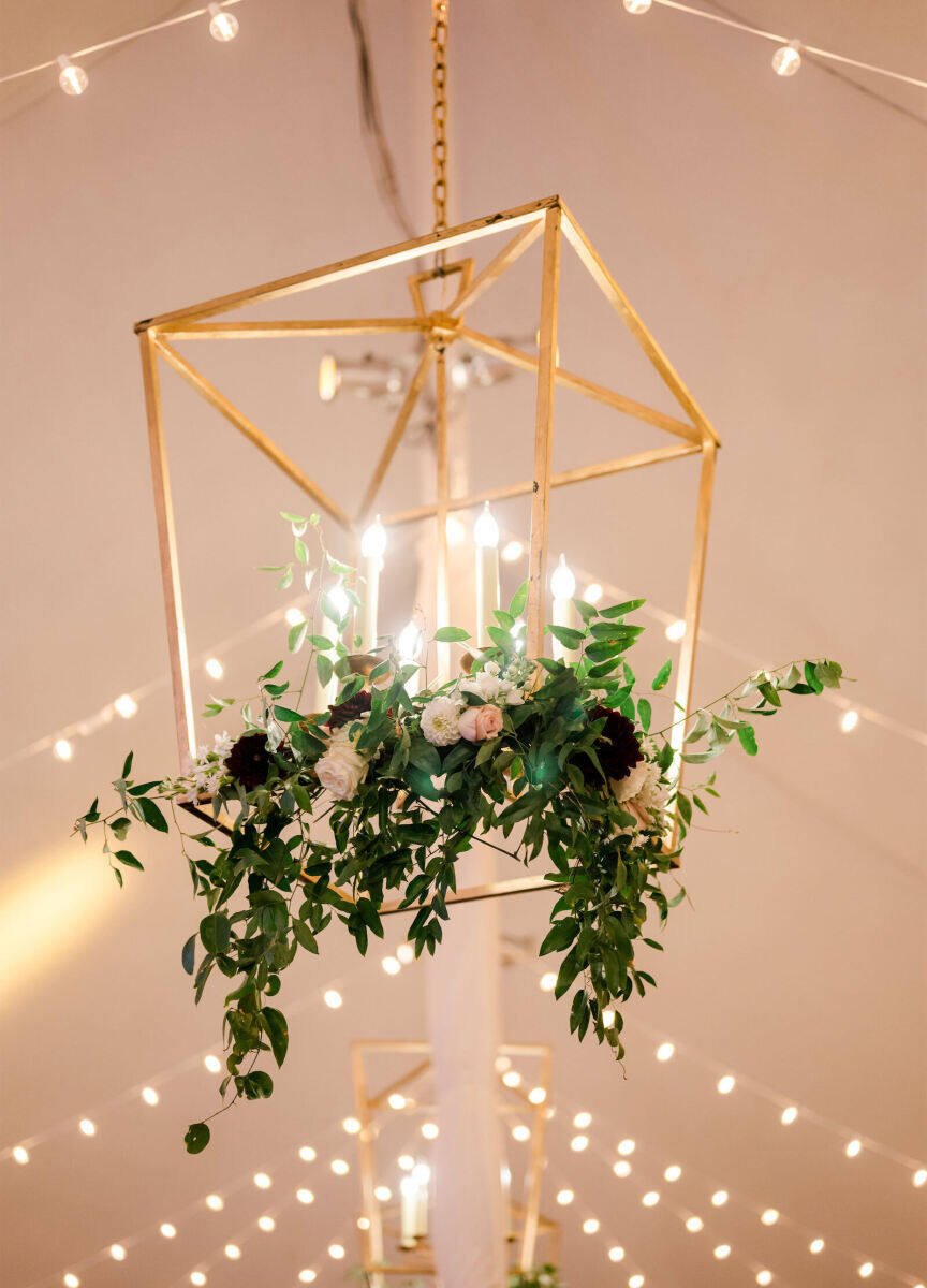 A gold lantern is decorated with fresh greenery and blooms in the tent of an elevated garden wedding reception.