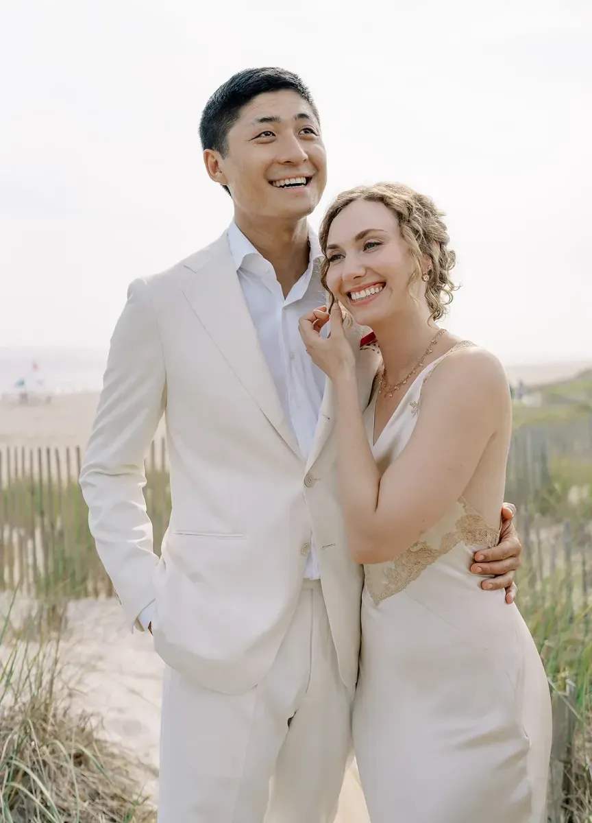 A bride and groom in cream-hued outfits embracing and smiling on the beach in Water Mill, New York