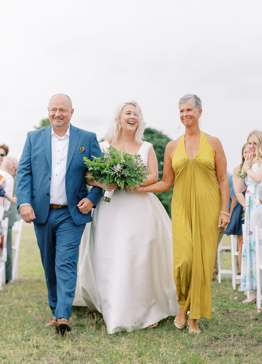 A bride walks down the aisle with her father and mother during her enchanted waterfront wedding.