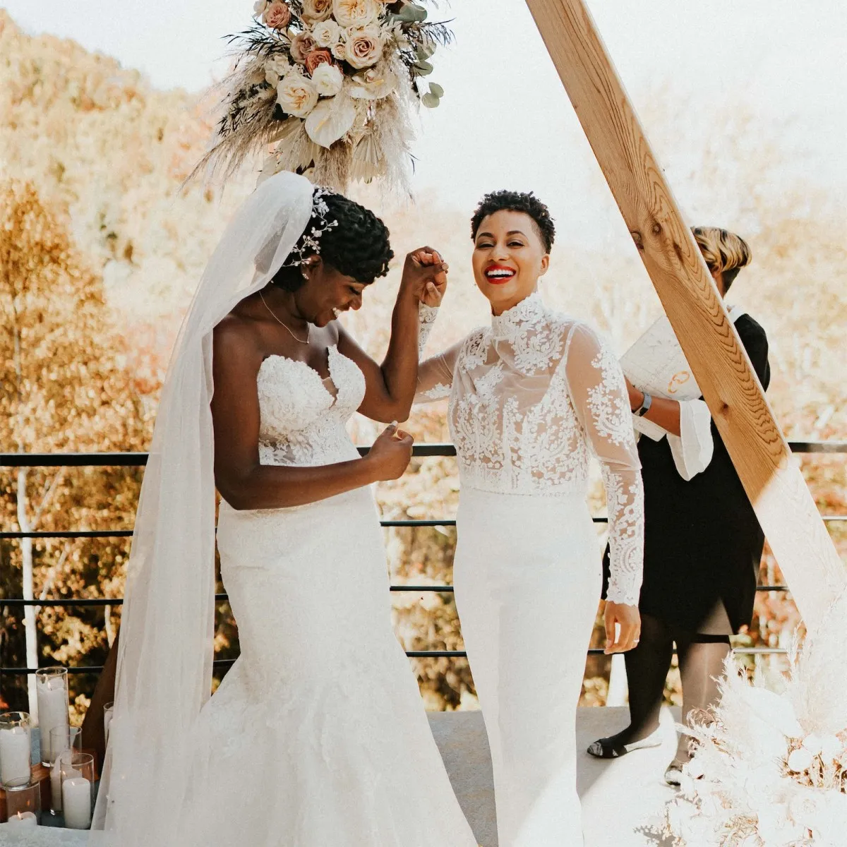 A wedding couple holding hands and smiling at the altar, with fall trees in the background.