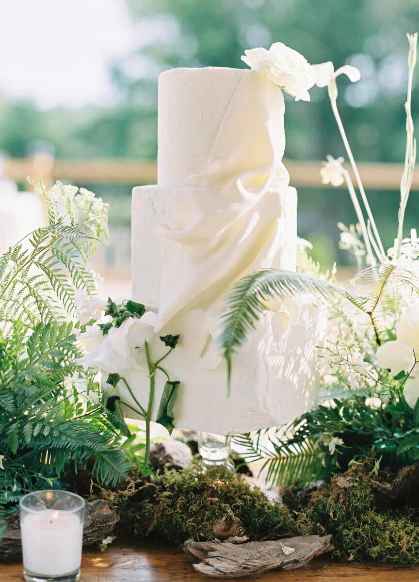 A asymmetrical wedding cake surrounded by ferns and moss and on display at a forest wedding in Canada.