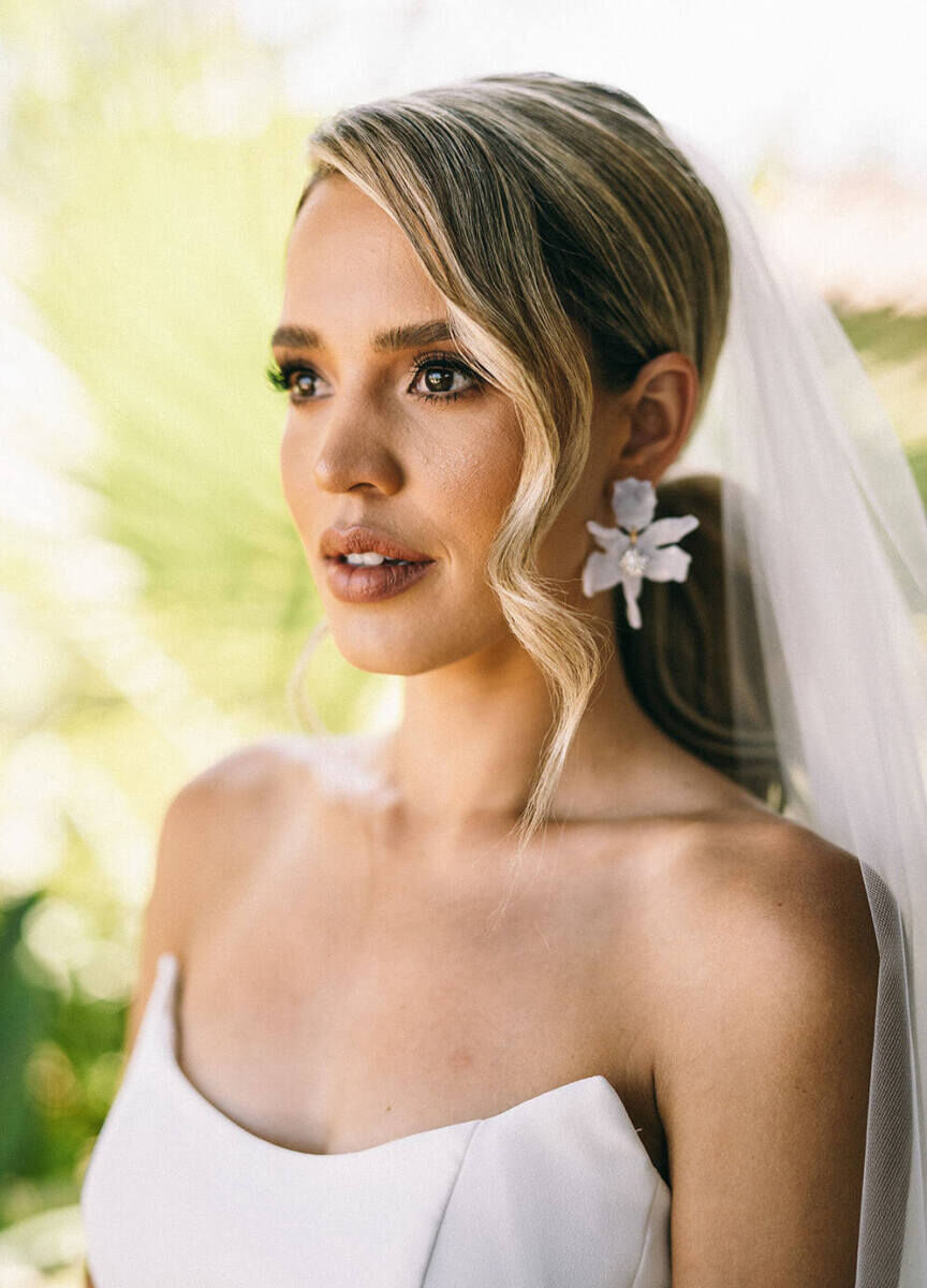 A portrait of a bride wearing a floral earring, strapless dress, and soft waves in her hair on the morning of her glam beach wedding.