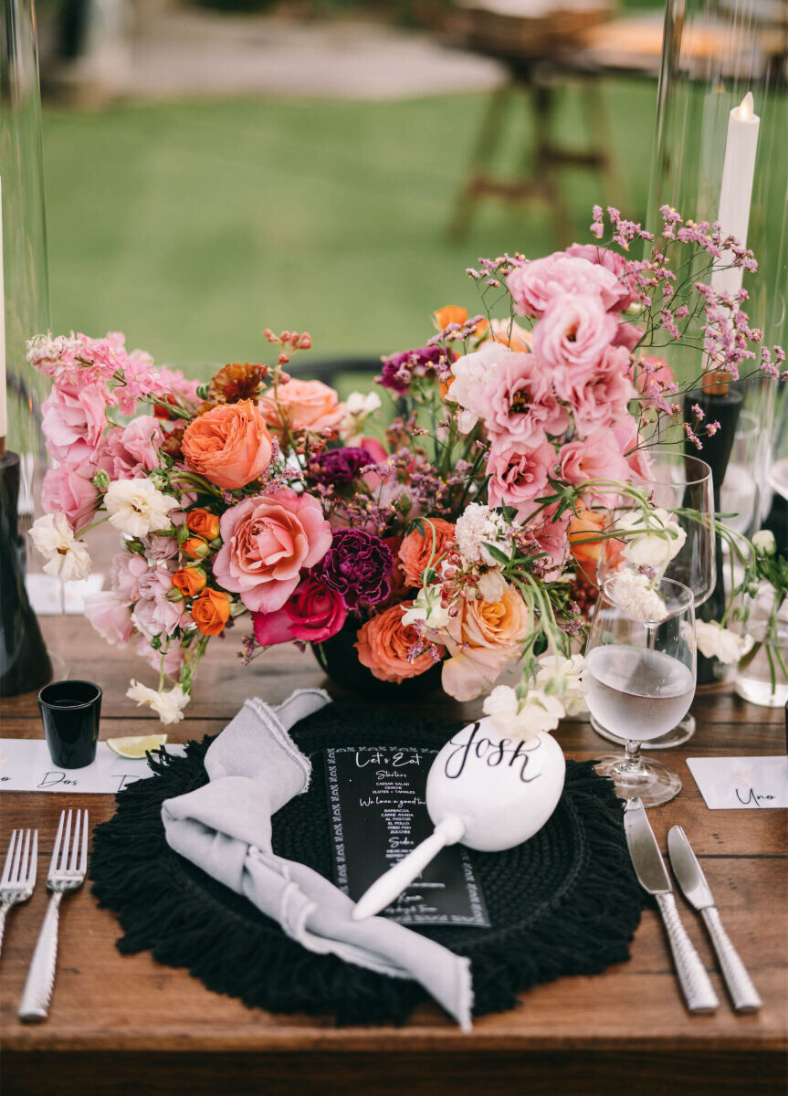 A pink and orange arrangement of flowers popped against the black fringe chargers and white maracas at each place setting of this glam beach wedding reception.