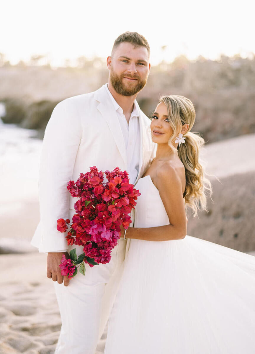 A bride holds her hot pink bougainvillea bouquet while posing for a portrait with her groom, who also wore white, at their glam beach wedding.