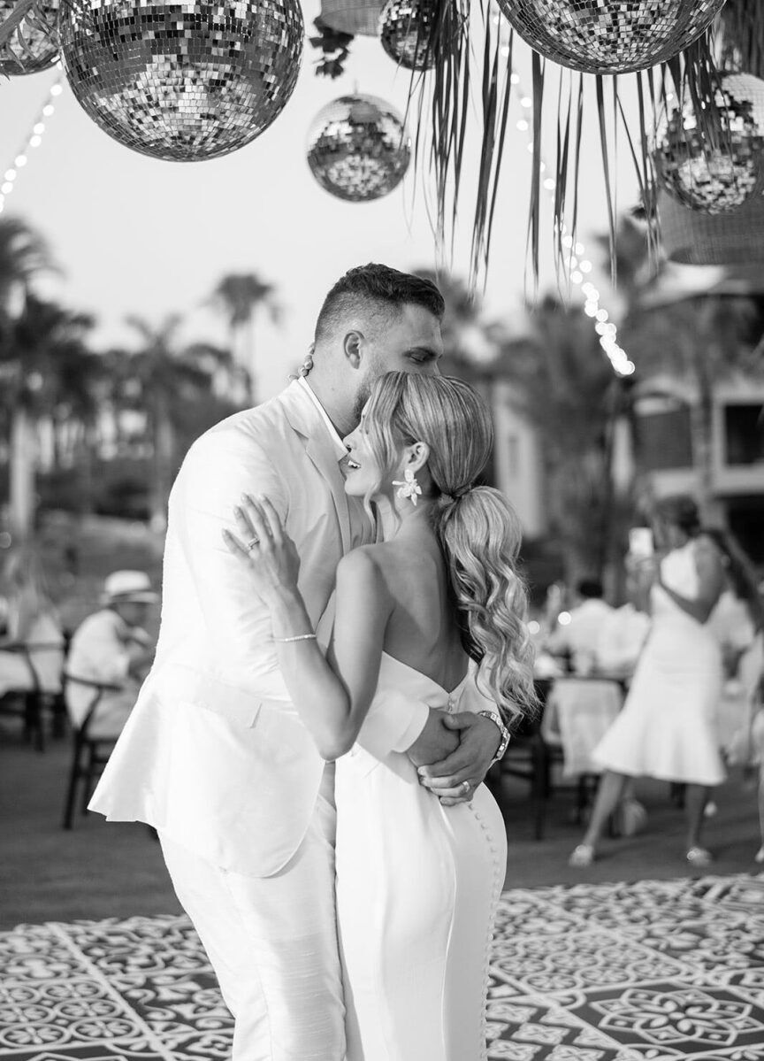 A pair of newlyweds, both wearing white, dance on a faux-tiled dance floor under an installation of disco balls during their glam beach wedding reception.