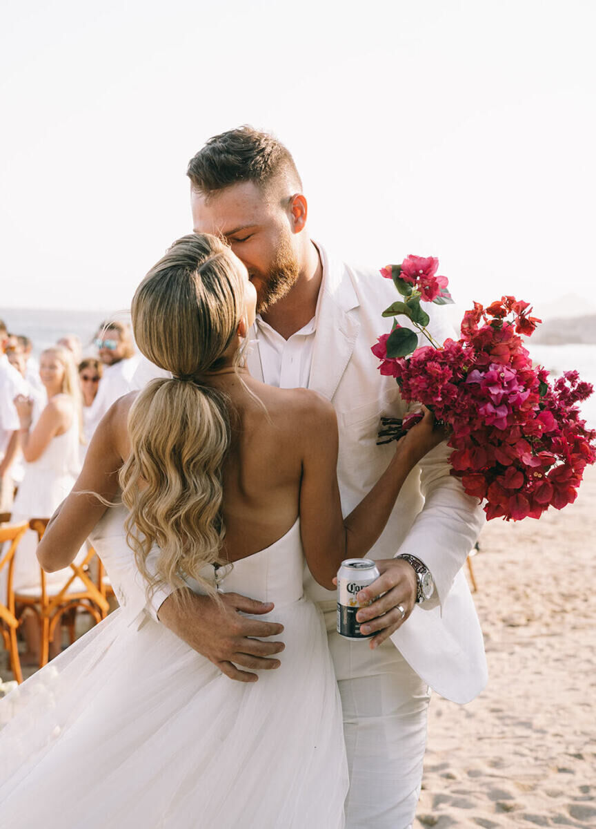 A groom grabs his wife and a beer at the end of the aisle following their glam beach wedding ceremony.