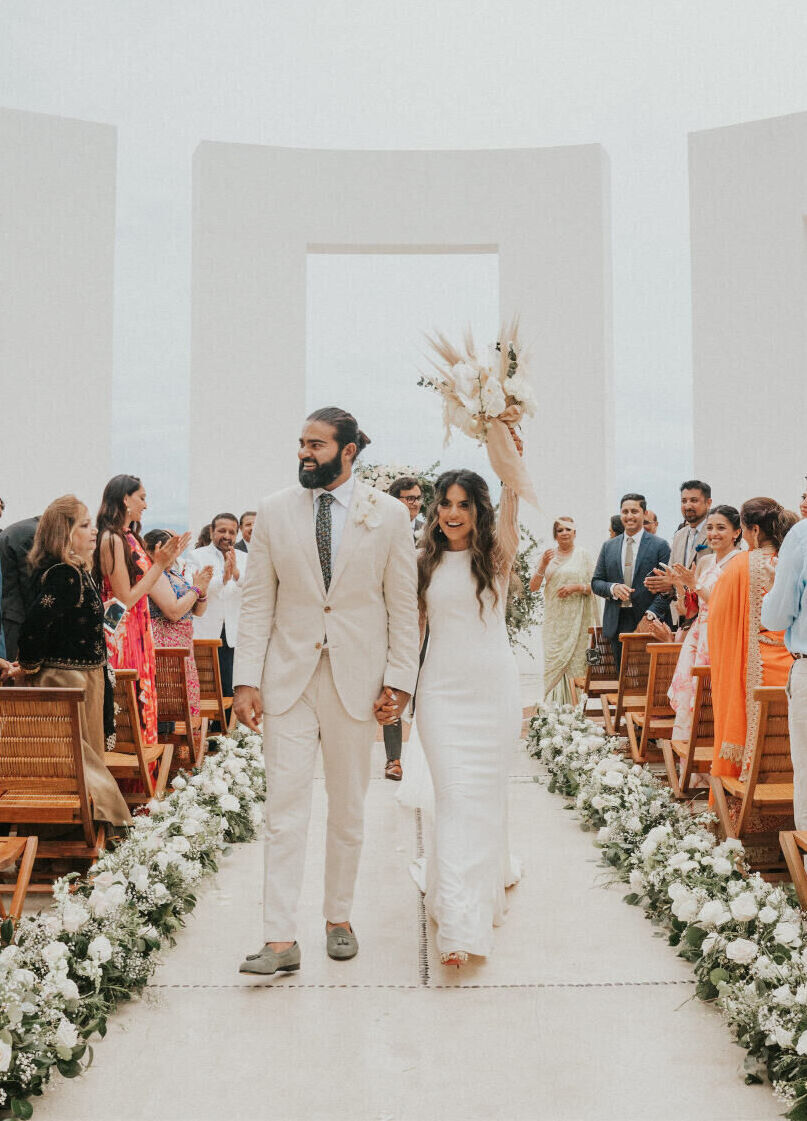 Grand Velas Riviera Nayarit: A bride and groom exiting a modern gazebo decorated with wooden chairs and white floral arrangements.