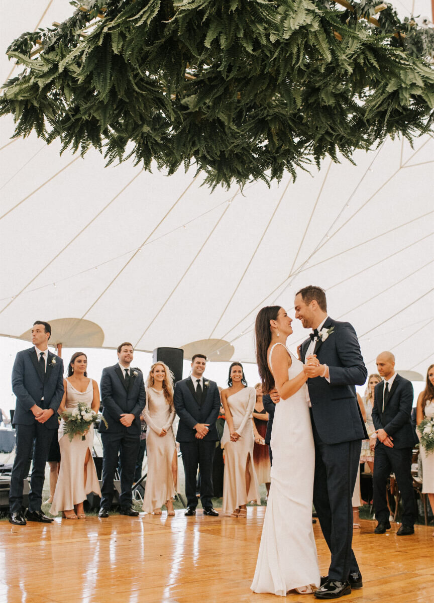 Holiday Wedding: A bride and groom slow dancing in the middle of a dance floor with greenery hanging overhead and guests looking at them in the background.