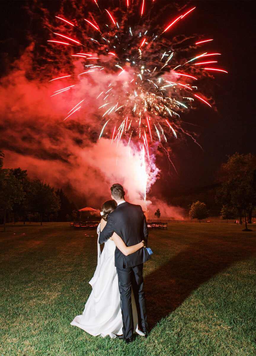 Holiday Wedding: Bride and groom with their arms around one another looking at fireworks in the distance