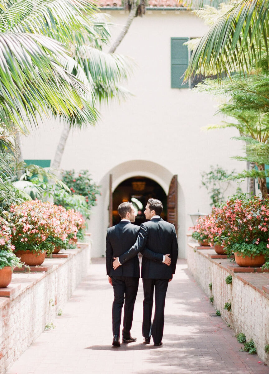 Holiday Wedding: Two grooms walking arm and arm down an outdoor path lined with palm trees.