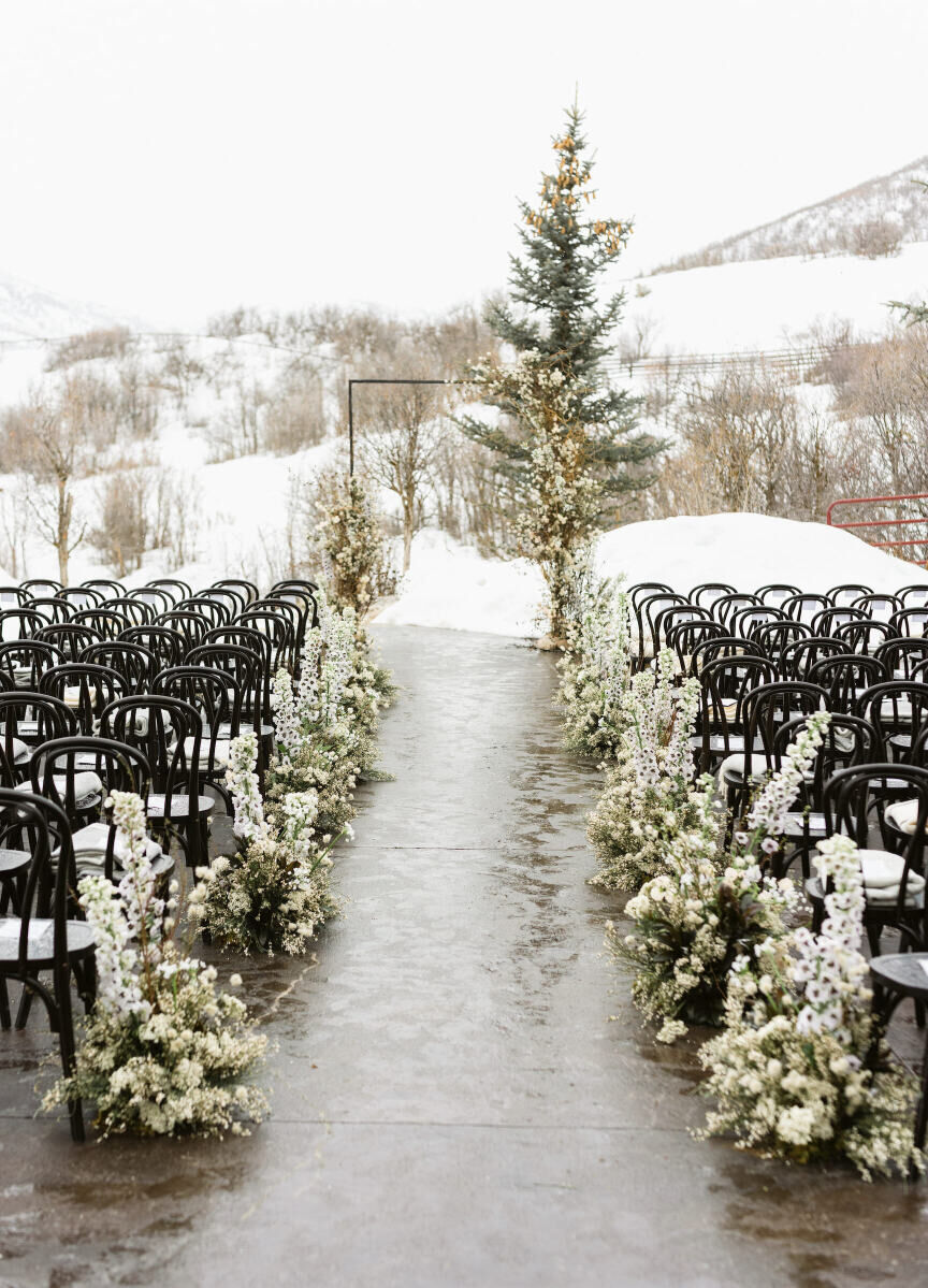 Holiday Wedding: A snowy outdoor ceremony set-up with mountains and a pine tree in the background. There are black chairs and white and green florals lining the aisle.